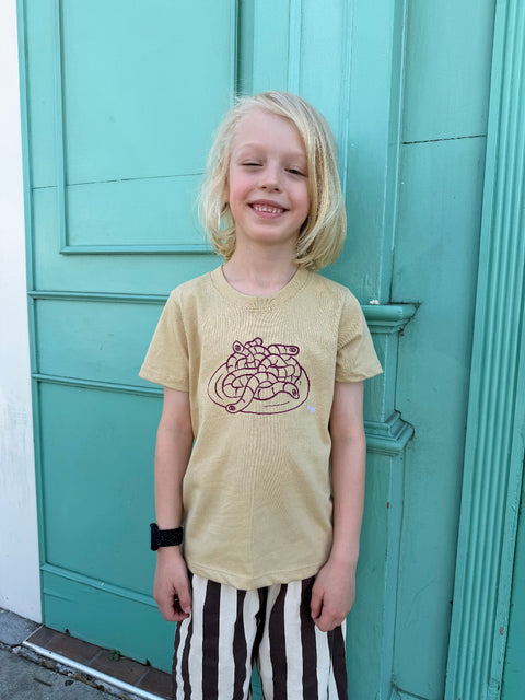 Child wearing a beige t-shirt with a graphic design in front of a teal door.
