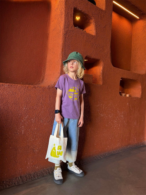 Child wearing a purple shirt and green hat standing against a textured brown wall.